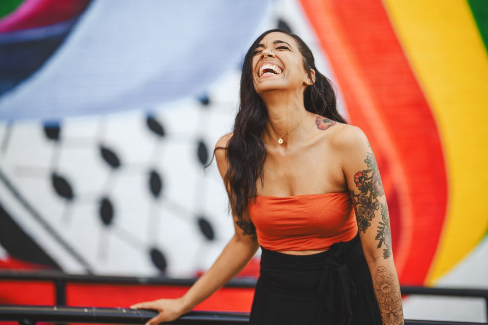 Person laughing with long dark hair, wearing an orange top and black pants, standing in front of a colorful mural. Person laughing with long dark hair, wearing an orange top and black pants, standing in front of a colorful mural.