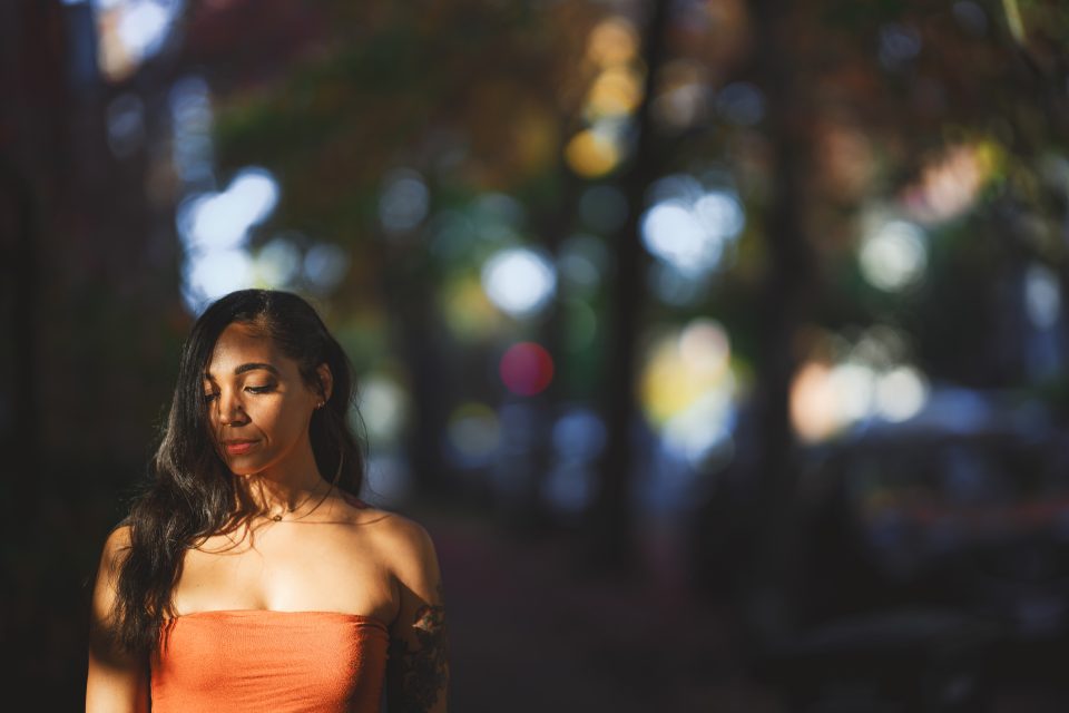 Woman in an orange top stands outdoors with eyes closed, surrounded by blurred trees and soft light. Woman in an orange top stands outdoors with eyes closed, surrounded by blurred trees and soft light.