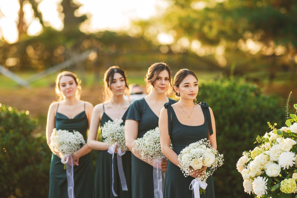 Four bridesmaids in dark green dresses holding white floral bouquets stand outdoors in a line. Four bridesmaids in dark green dresses holding white floral bouquets stand outdoors in a line.