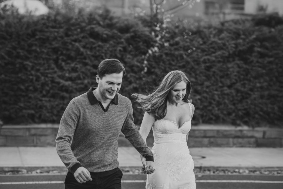 A couple holding hands and walking outdoors, both smiling. The woman is wearing a strapless dress, and the man is in a sweater. The background features shrubs and a sidewalk. A couple holding hands and walking outdoors, both smiling. The woman is wearing a strapless dress, and the man is in a sweater. The background features shrubs and a sidewalk.