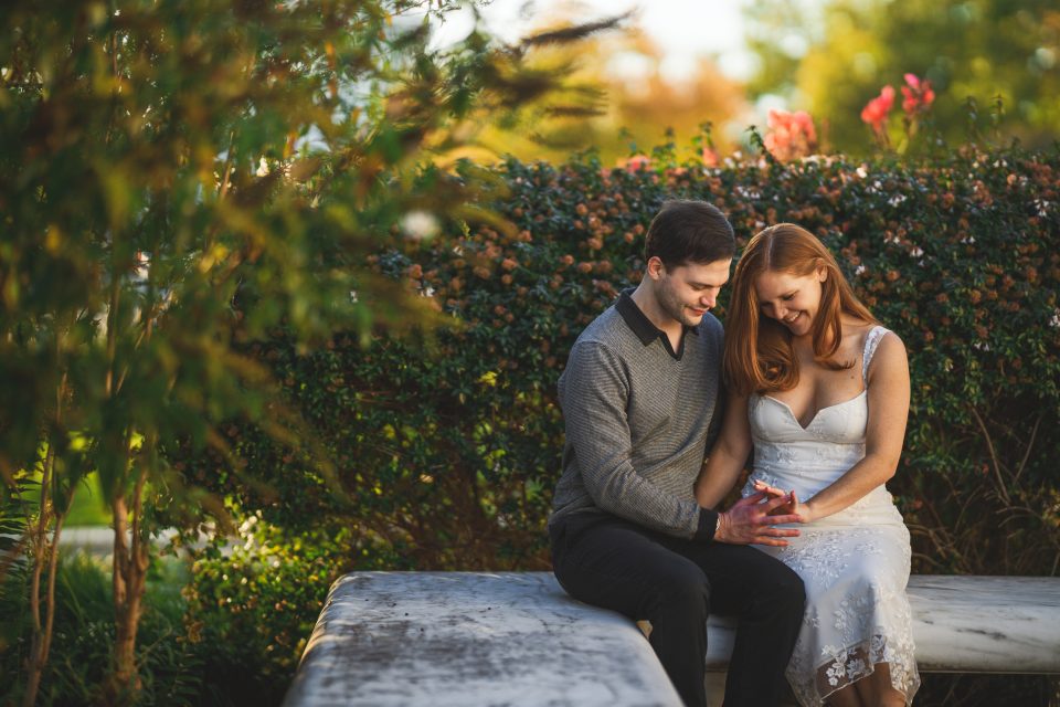 A couple sits on a stone bench in a garden, smiling and holding hands. The woman wears a white dress, and the man wears a grey sweater. A couple sits on a stone bench in a garden, smiling and holding hands. The woman wears a white dress, and the man wears a grey sweater.