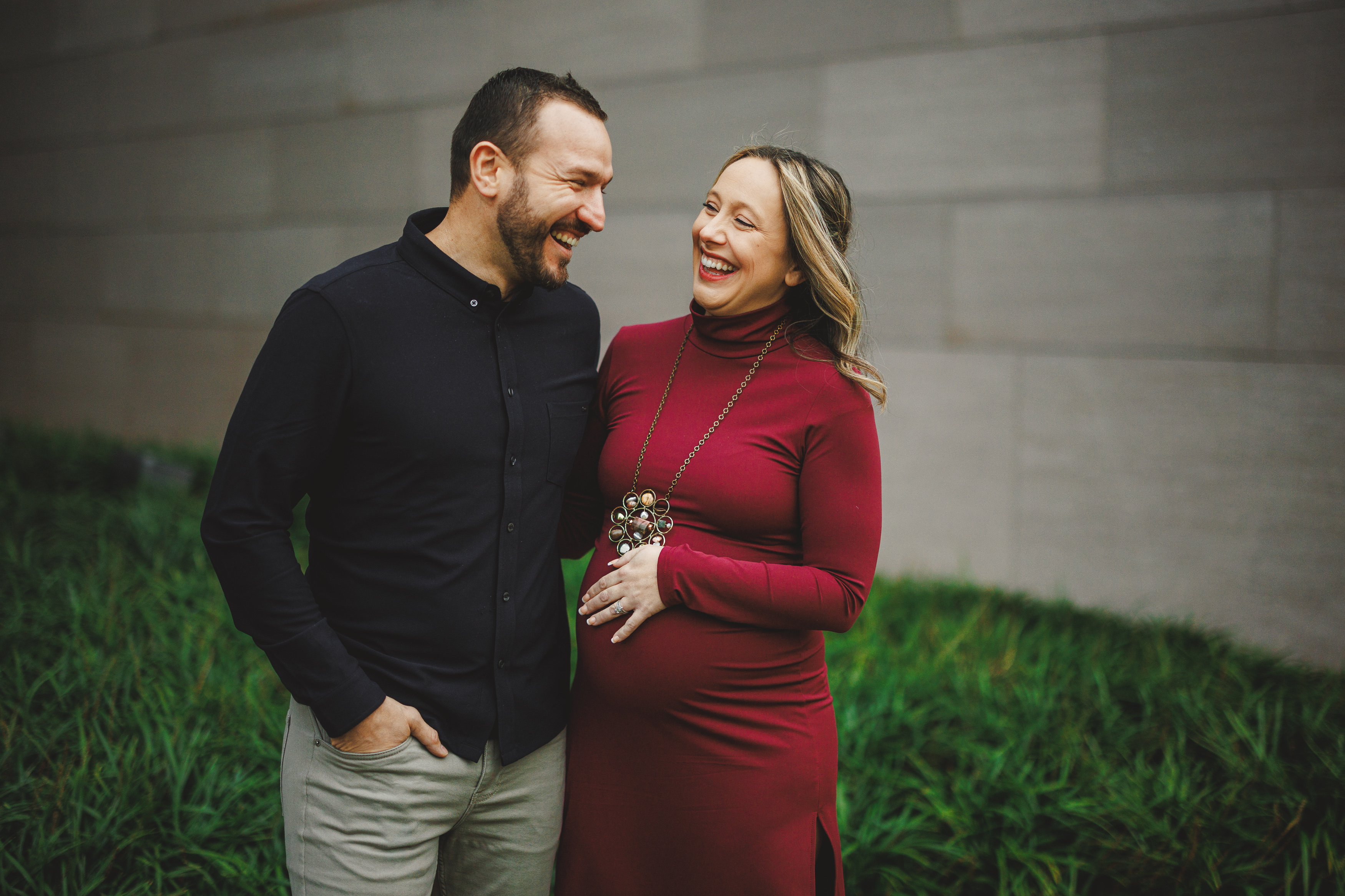 A smiling couple stands outdoors. The woman in a red dress is pregnant and holding her belly, while the man in a black shirt has his hand in his pocket. They are near a gray wall and greenery.