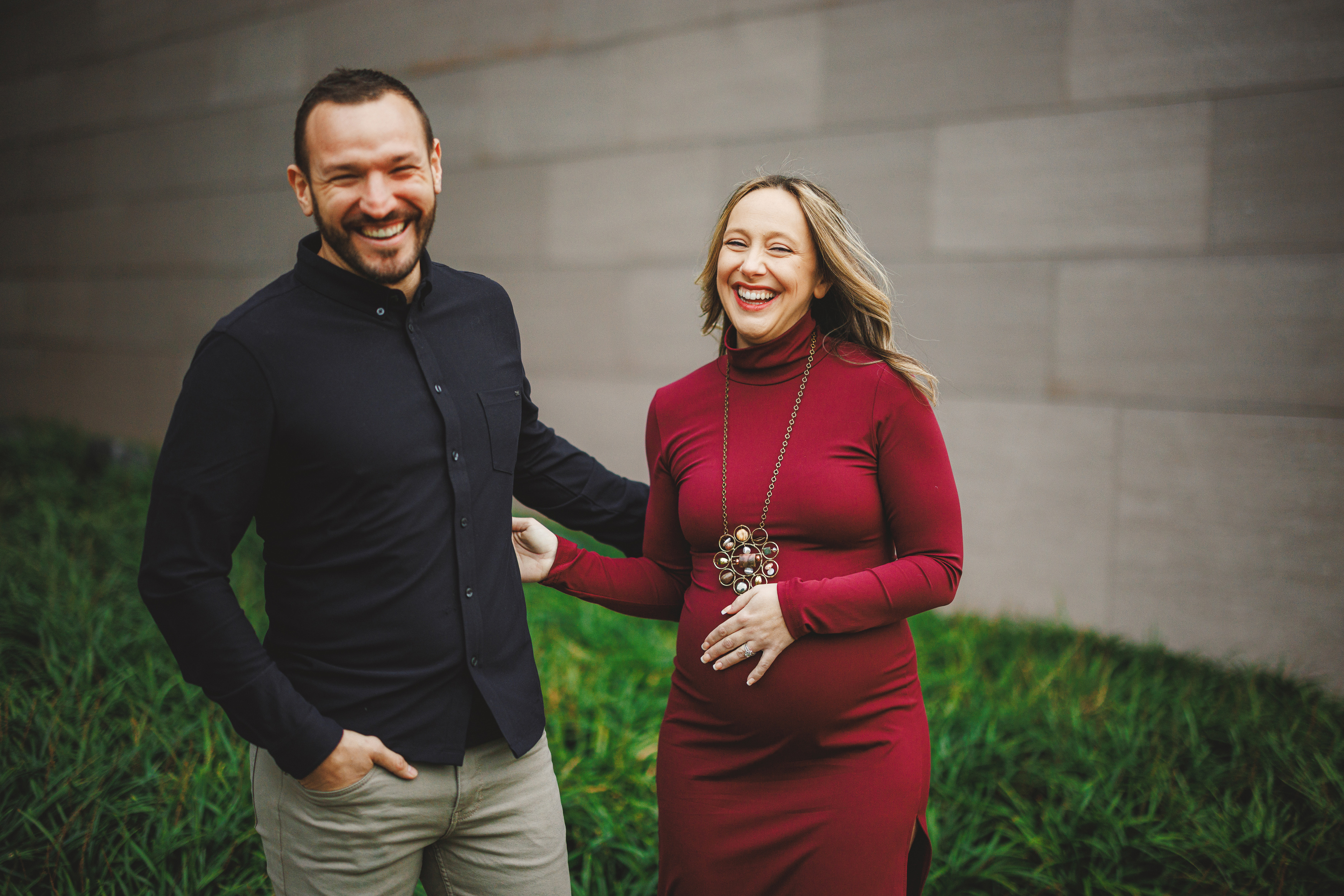 A man and a pregnant woman stand smiling outdoors, with the woman wearing a red dress and the man in a black shirt.