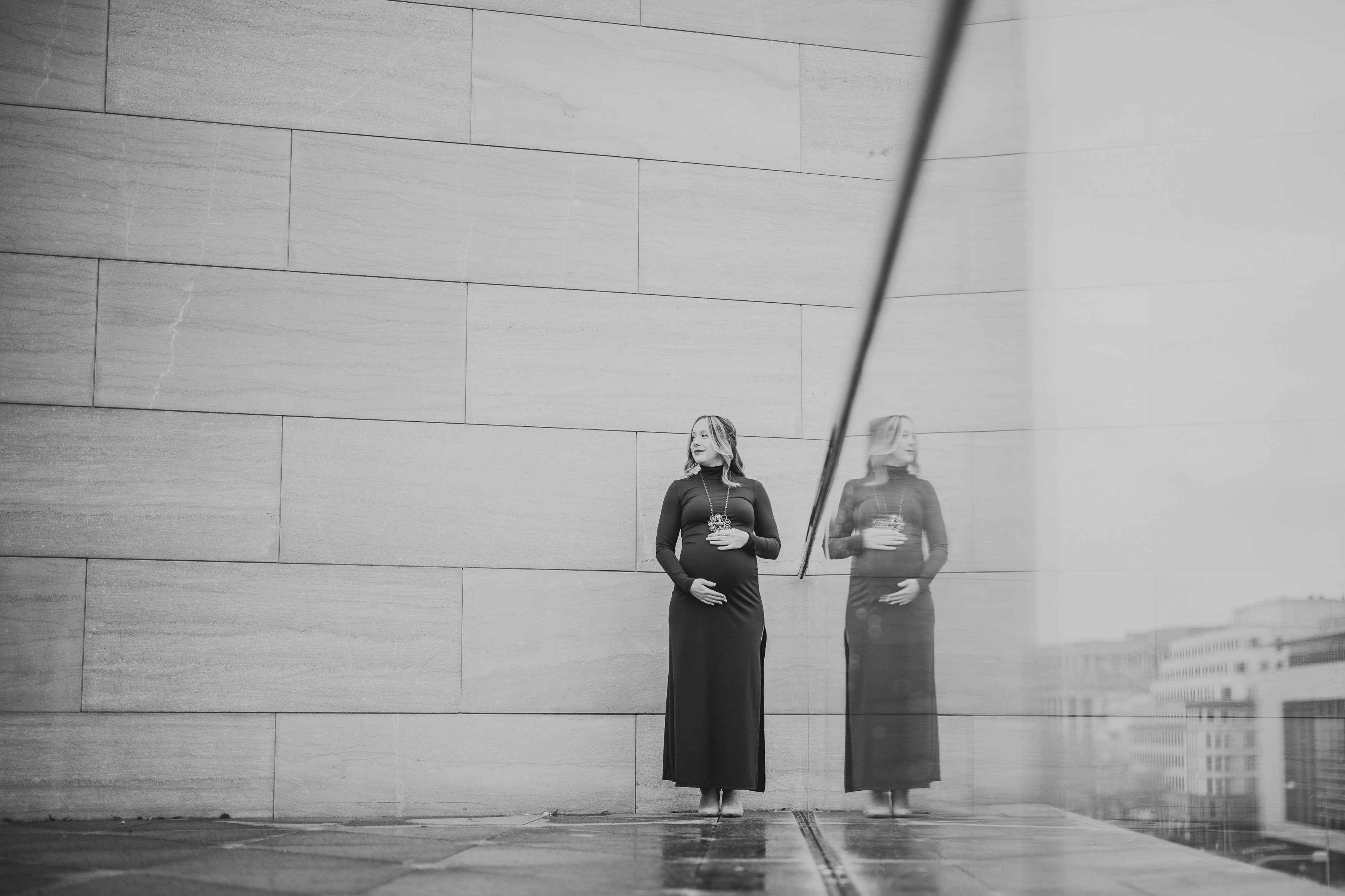 A woman in a long dress stands next to a reflective glass wall, holding her stomach. The background features large stone tiles.
