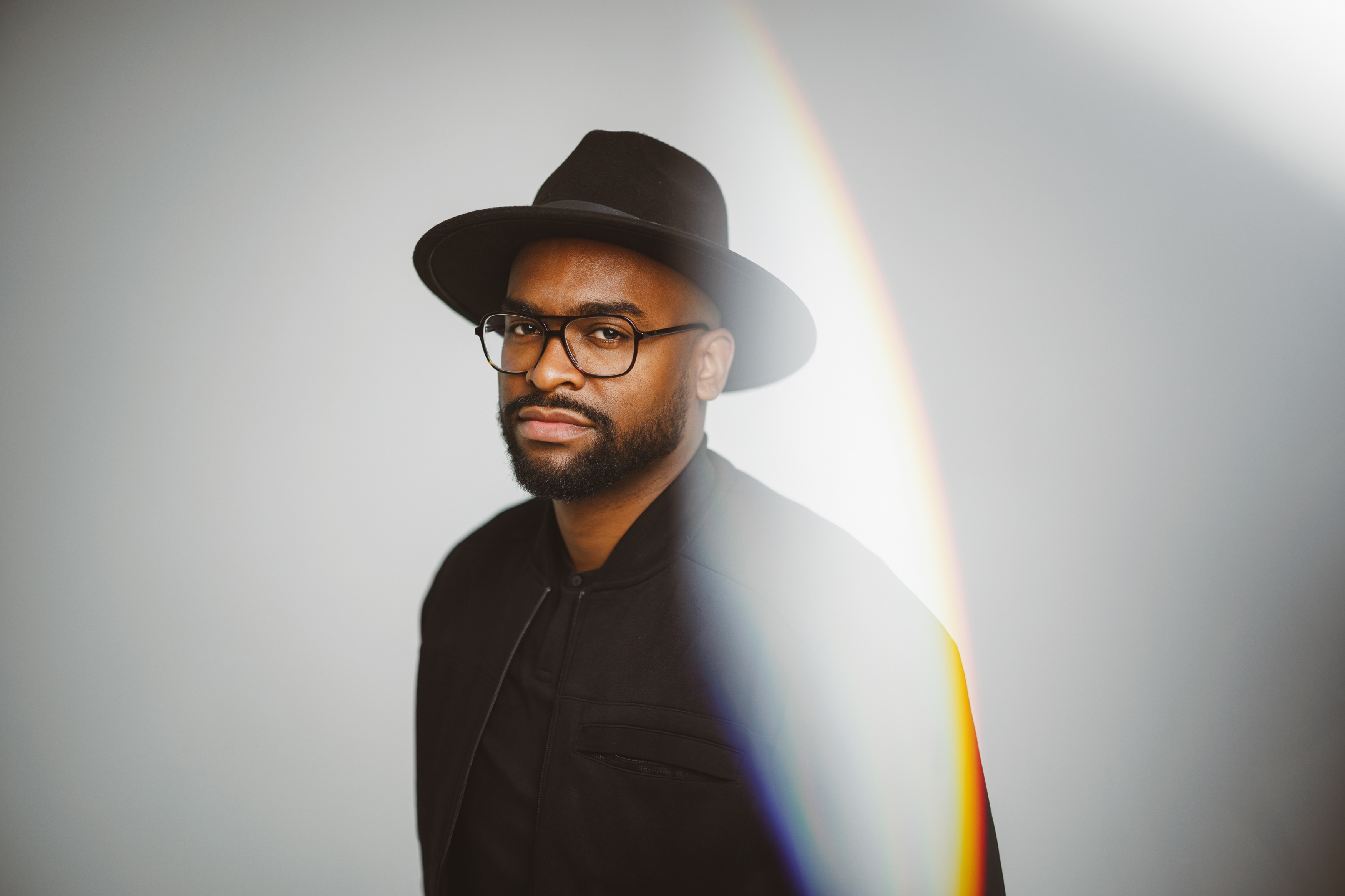 A man wearing glasses and a wide-brimmed hat stands against a light background with a rainbow streak.