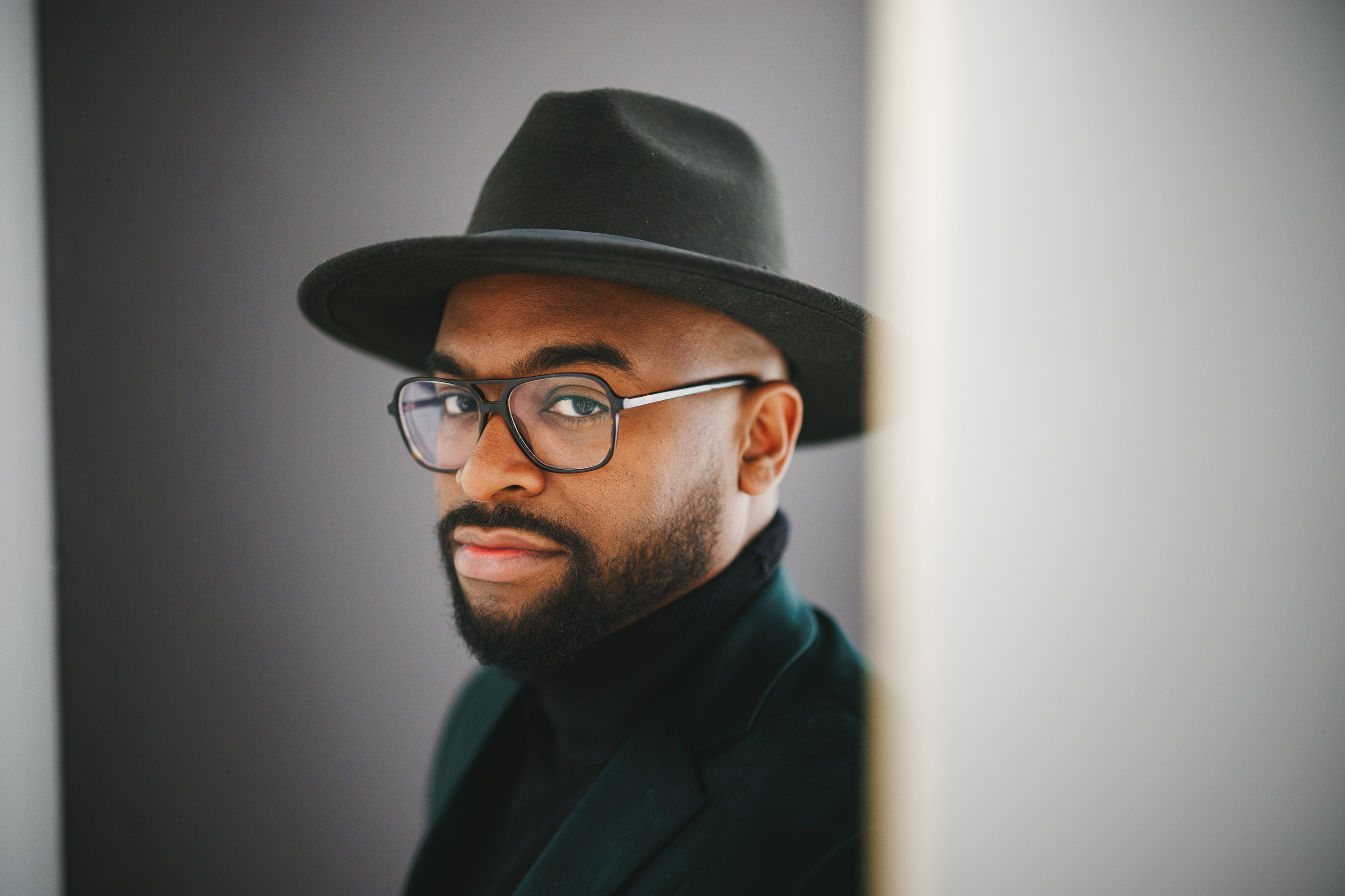 A man wearing a dark hat, glasses, and a black shirt appears to look at the camera against a neutral background.