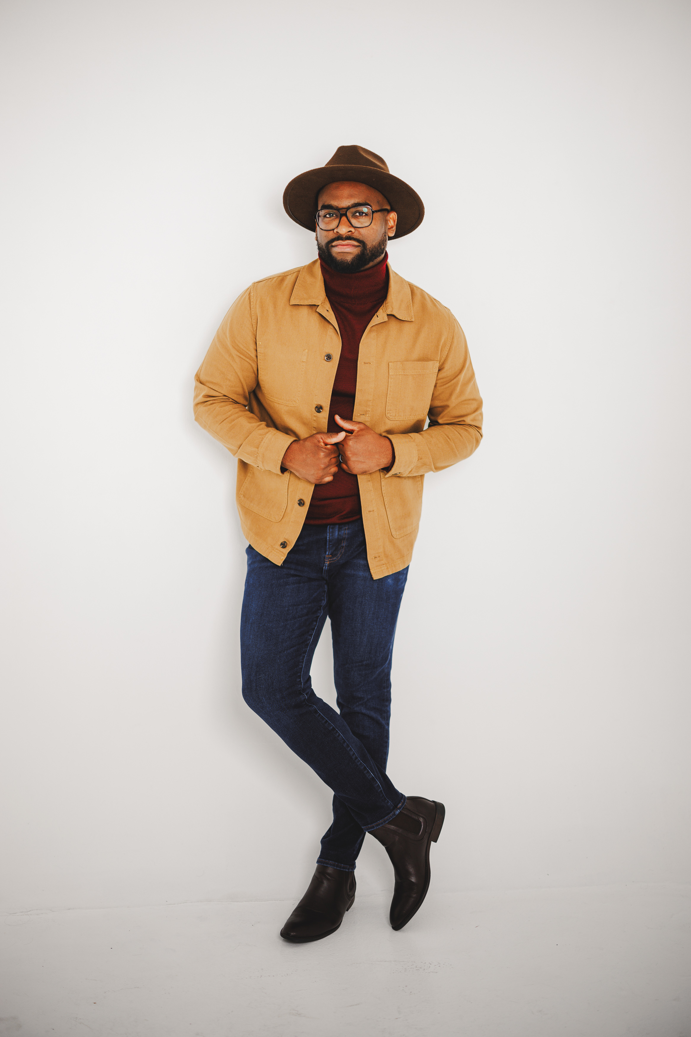 A person wearing a brown hat, tan jacket, red sweater, blue jeans, and black boots stands against a plain white background with arms crossed.