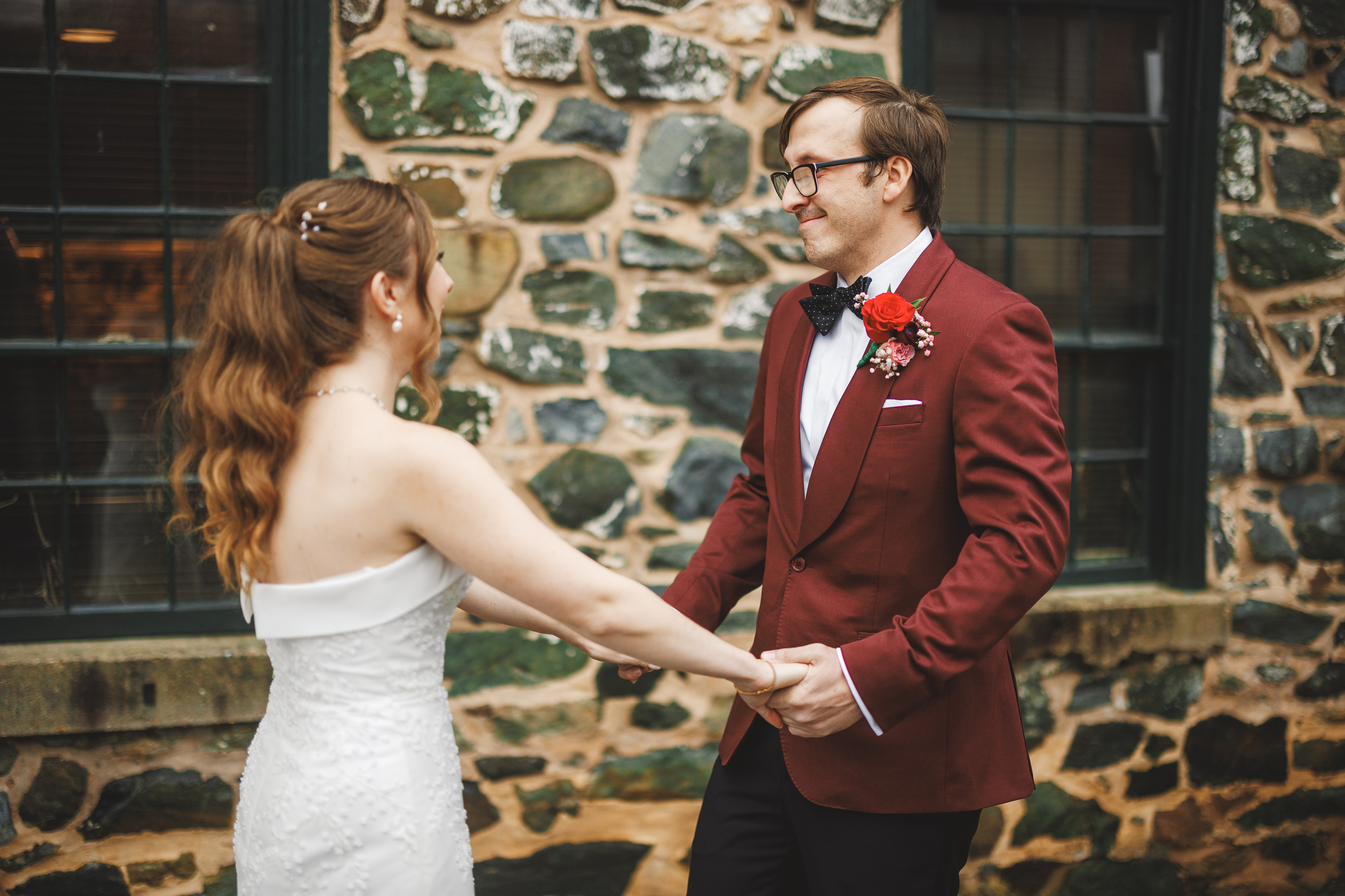 A couple holding hands, facing each other, in formal attire. The man wears a red suit with a bow tie, and the woman is in a white dress. Stone wall in the background.