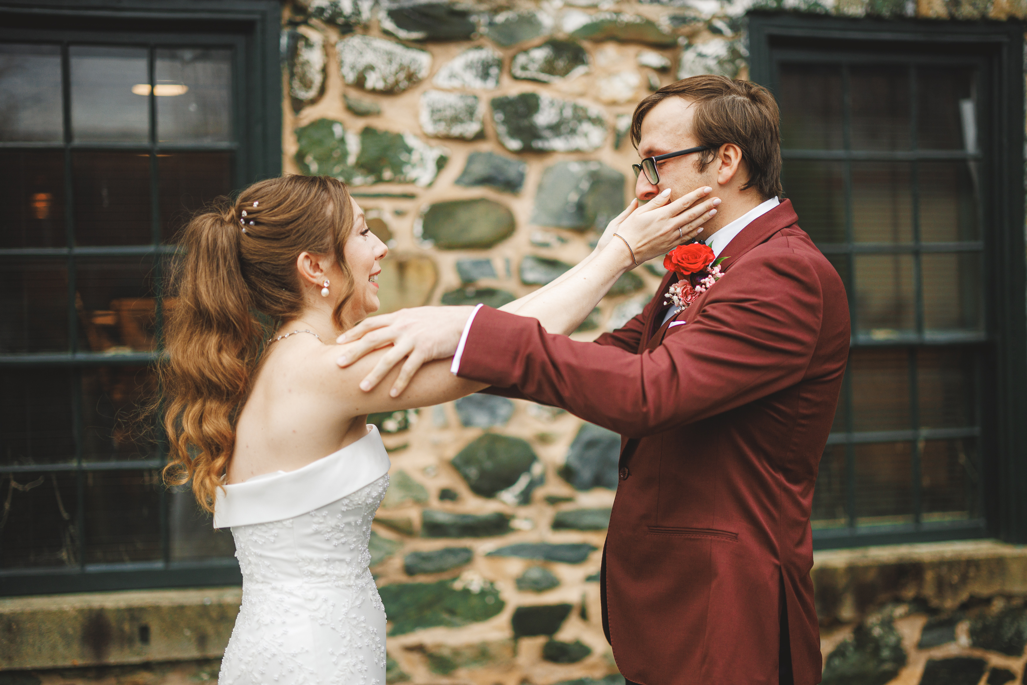 A bride and groom stand outside a stone building. The bride is touching the groom's face, and both are smiling. The groom wears a red suit with a red boutonniere.
