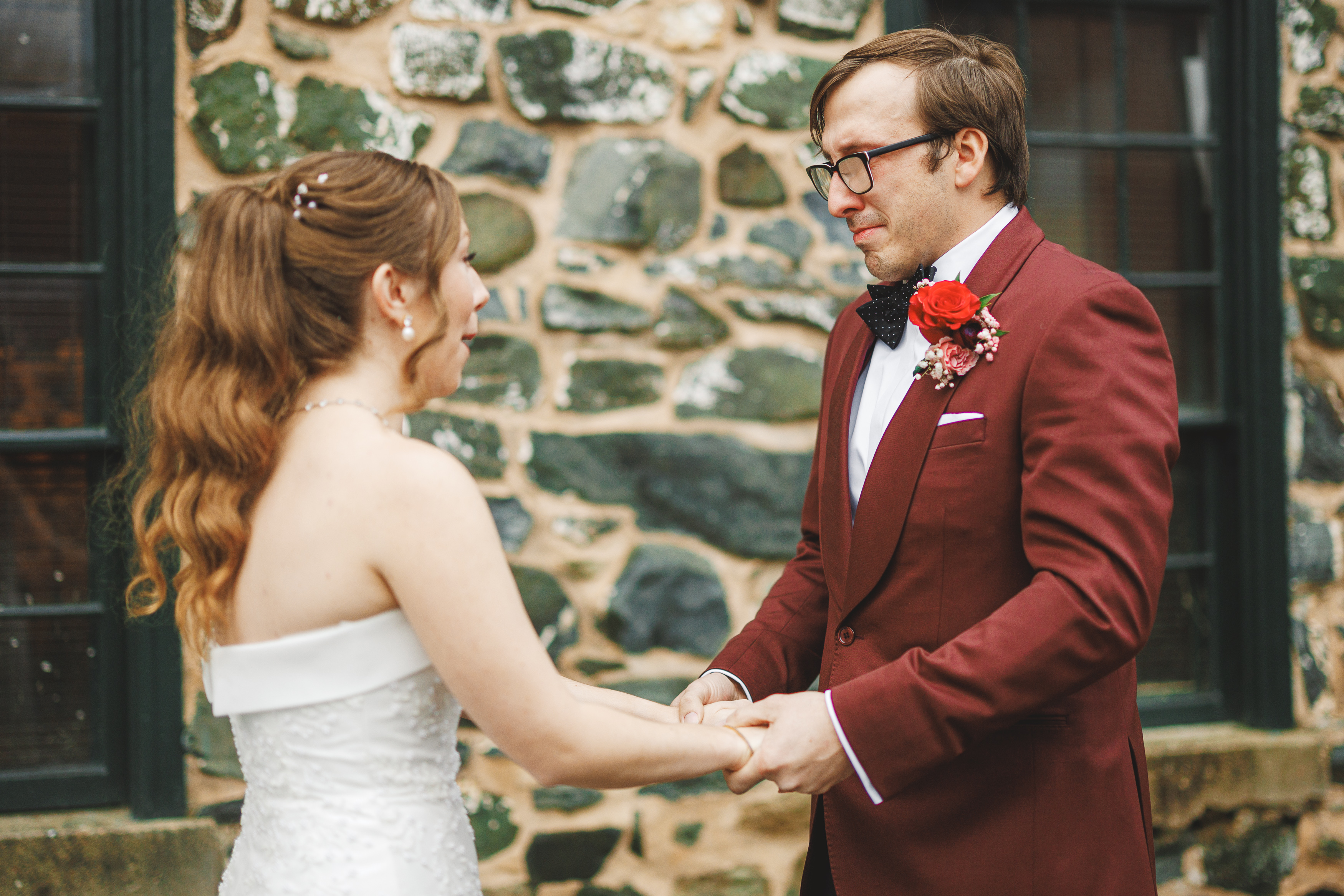 A bride and groom stand holding hands, facing each other. The groom wears a burgundy suit with a red rose boutonniere, while the bride is in a white dress. They are in front of a stone wall.