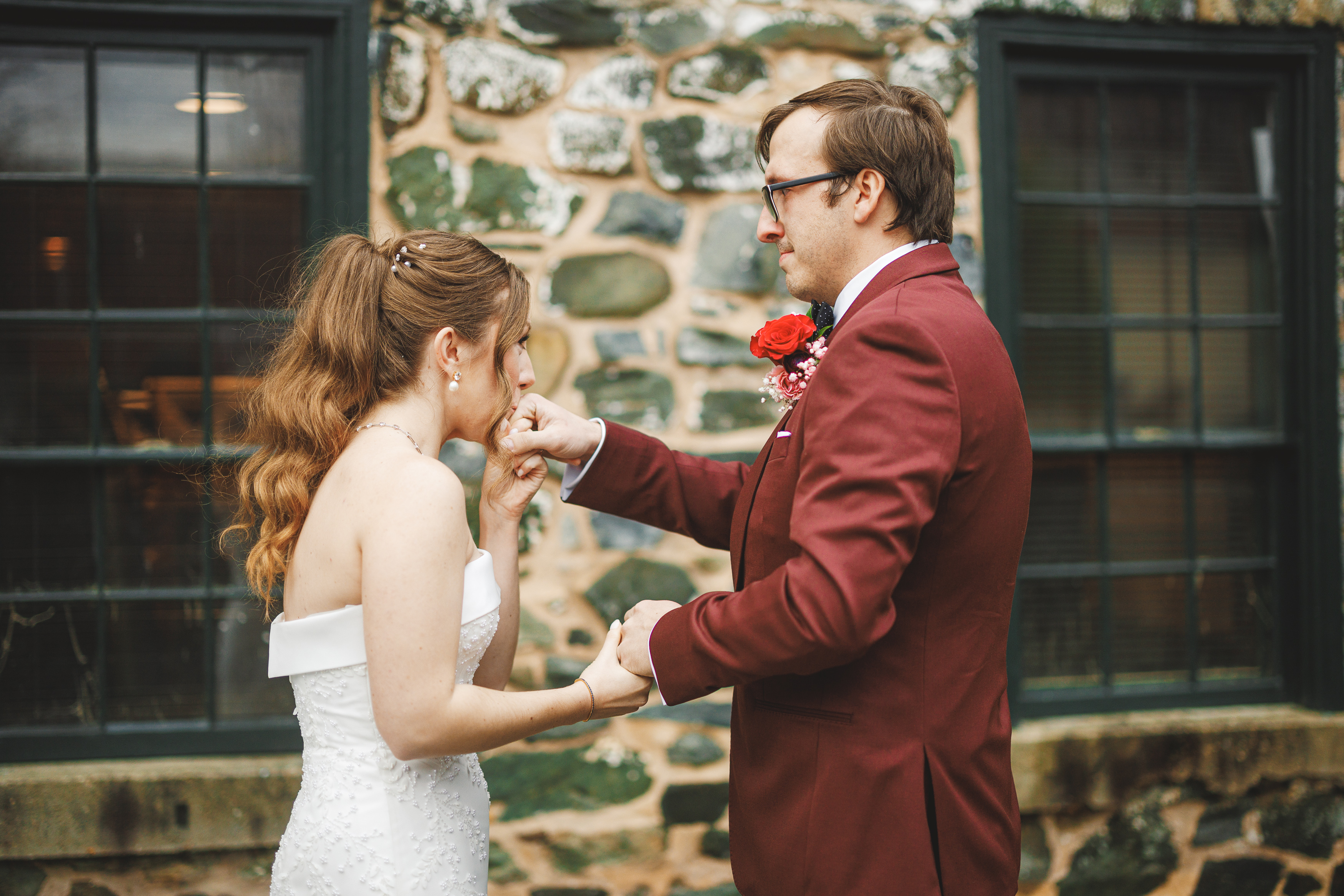 A bride and groom stand outside, surrounded by stone walls. The groom, in a maroon suit, kisses the bride's hand. The bride wears an off-shoulder white gown. Both appear emotional.