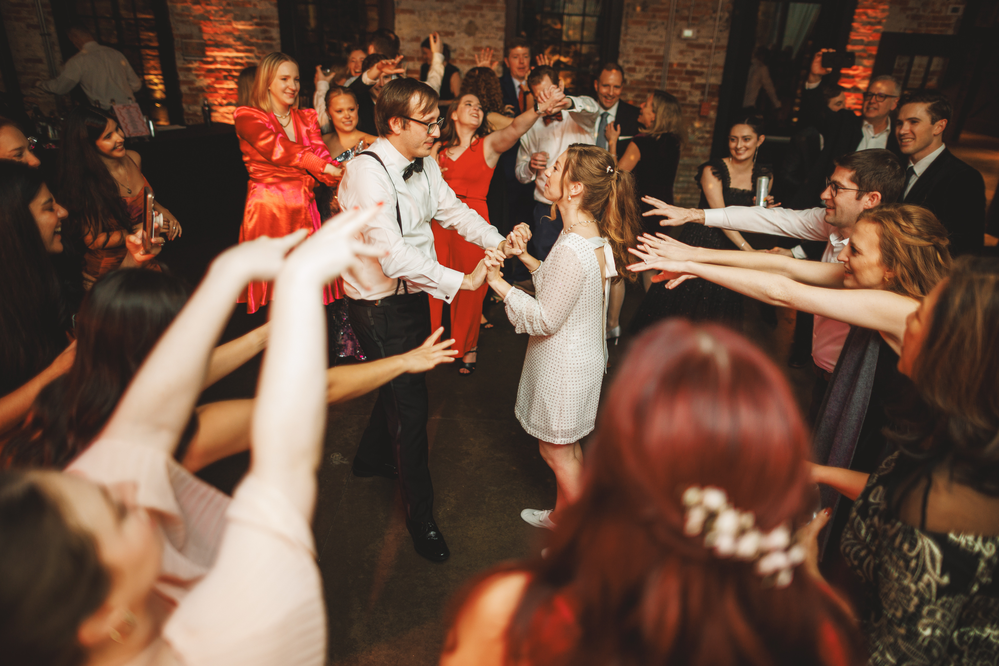 A group of people are joyfully dancing, with a couple in the center holding hands. The setting is a warmly lit indoor venue with exposed brick walls.