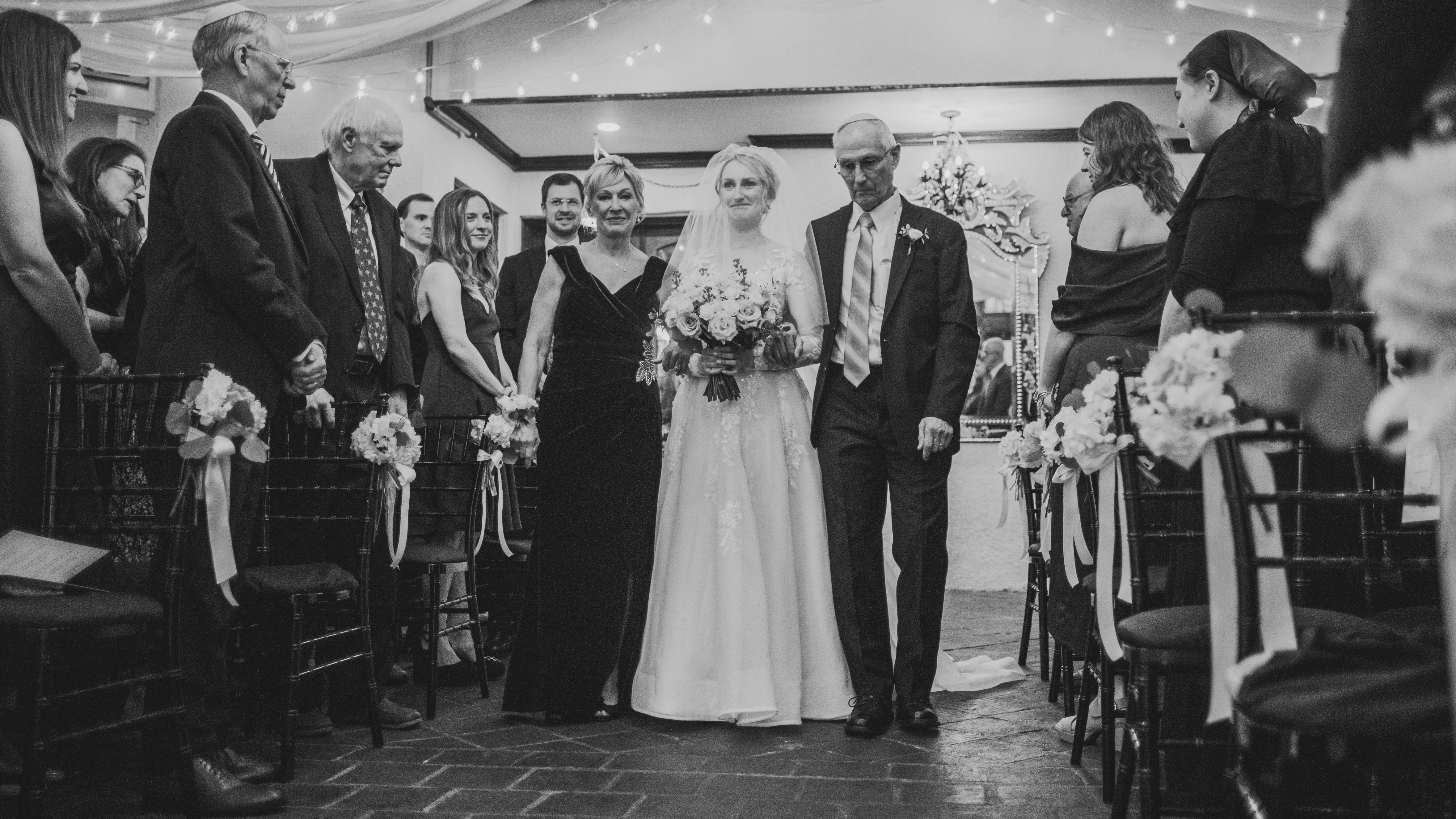 Bride in a white gown walking down the aisle with two escorts, surrounded by seated guests in a decorated venue.