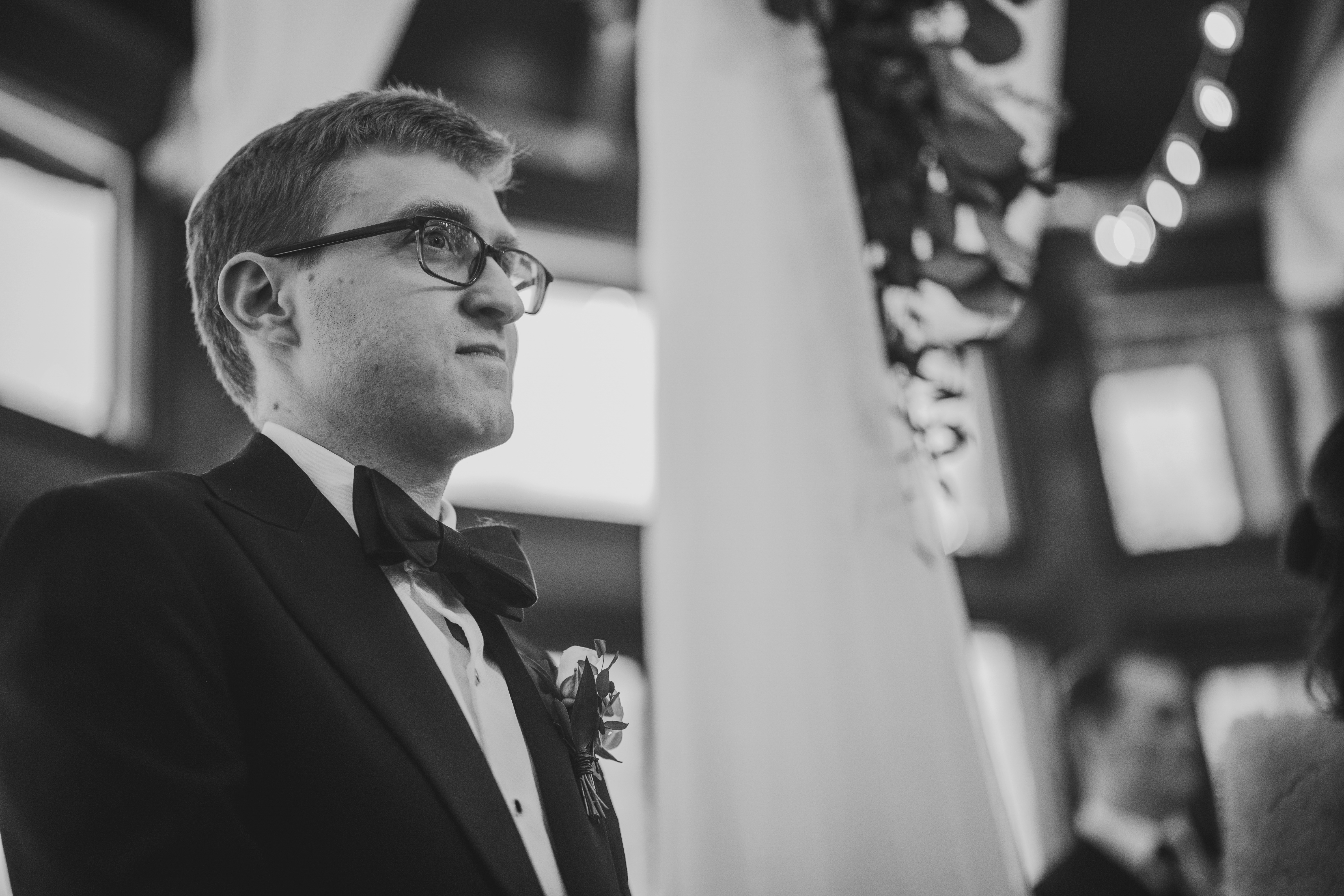 A man in a tuxedo and bow tie stands indoors next to a pillar, looking to his right.
