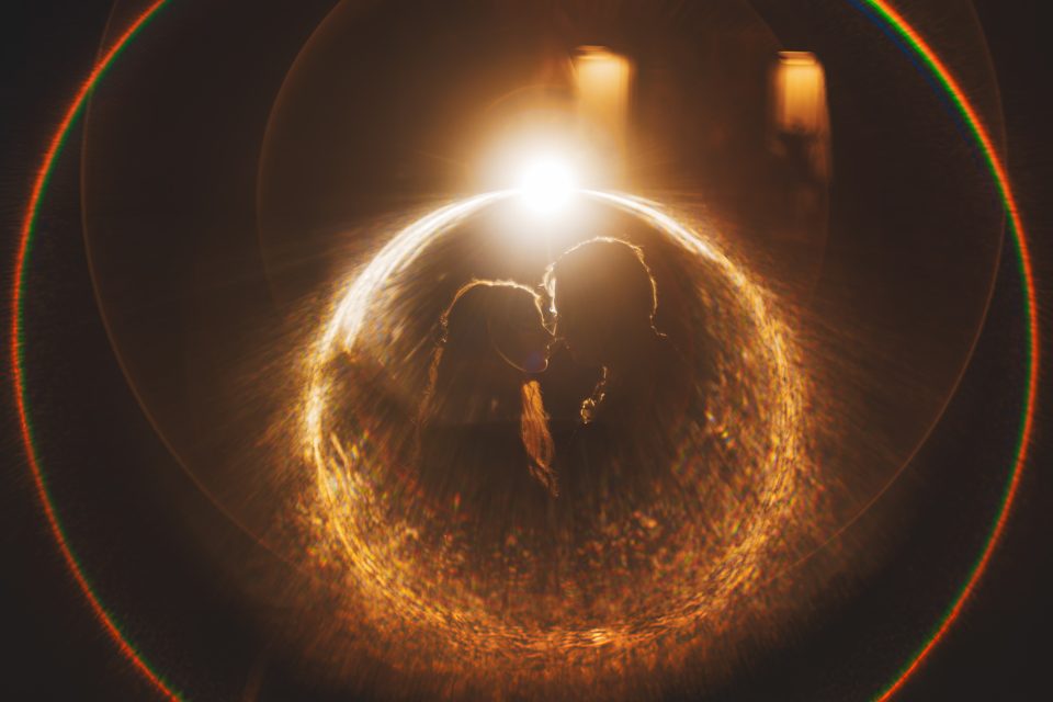 Silhouettes of two people facing each other with a bright light source behind them, creating a circular lens flare effect in warm tones.