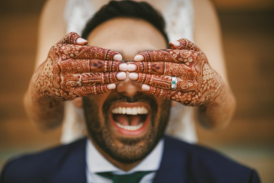 A smiling man in a suit is playfully covering his eyes with hands adorned with intricate henna designs and a ring.