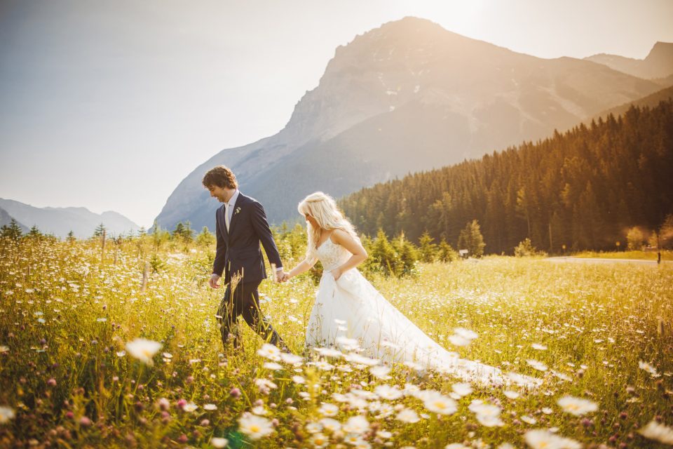 A bride and groom walk hand in hand through a sunlit field of wildflowers, with mountains and a forest in the background.