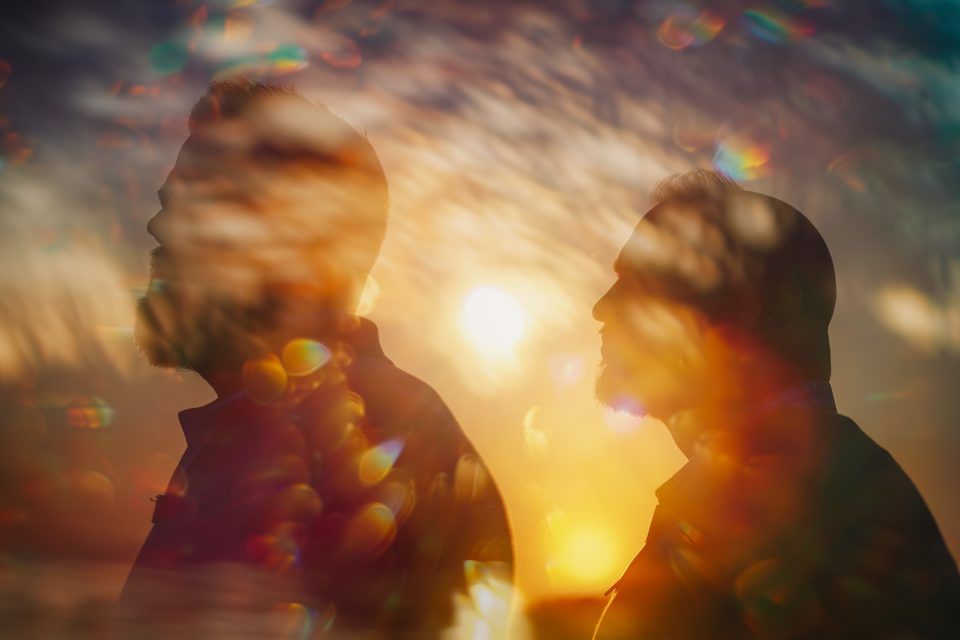 Two silhouetted people standing against a colorful, blurred, and abstract background with sunlight.