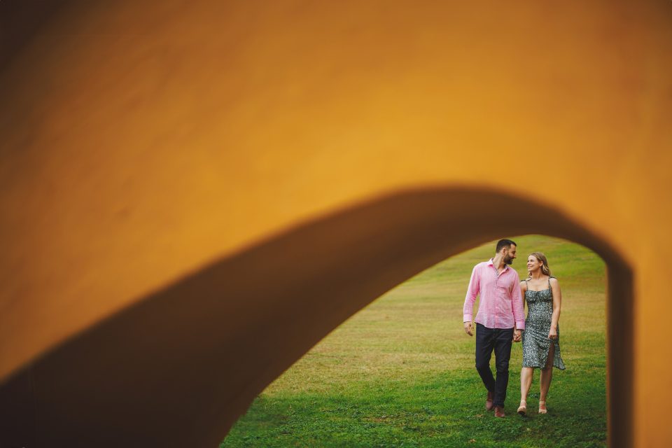A man and woman walk on grass under a large yellow arch, engaging in conversation.
