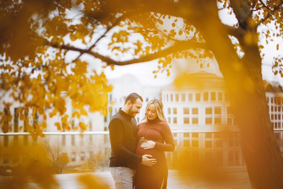 A couple stands closely under autumn trees with orange leaves, looking at the woman's pregnant belly, on an outdoor urban walkway.
