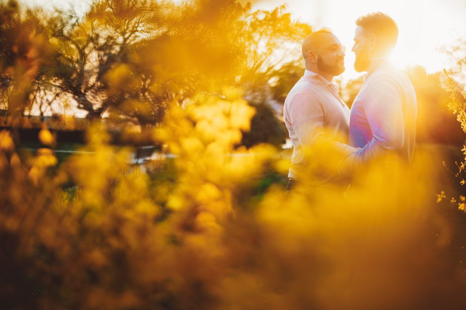 Two people stand close together in a park during sunset, surrounded by trees and yellow foliage, with sunlight creating a warm glow.