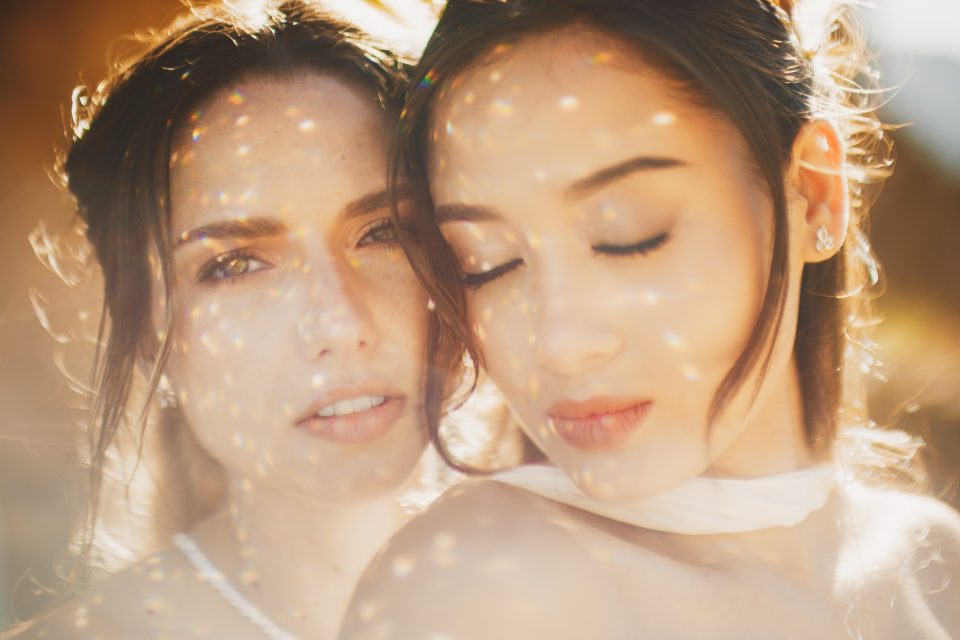 Two women with soft, warm lighting and bokeh effects on their faces. One is looking at the camera, while the other has her eyes closed.