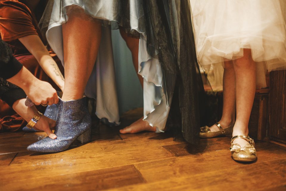 Person putting on blue glitter boot while surrounded by others in evening dresses. Another person stands nearby in gold shoes. Wooden floor visible.