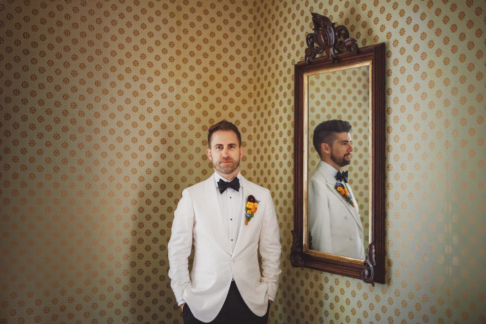A man in a white tuxedo with a black bow tie stands in front of a patterned wall. His reflection is visible in a framed mirror to his right.
