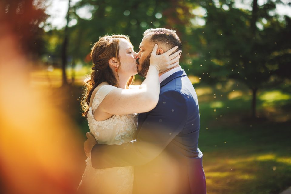 A couple kisses outdoors, surrounded by greenery and sunlight, with small particles visible in the air.