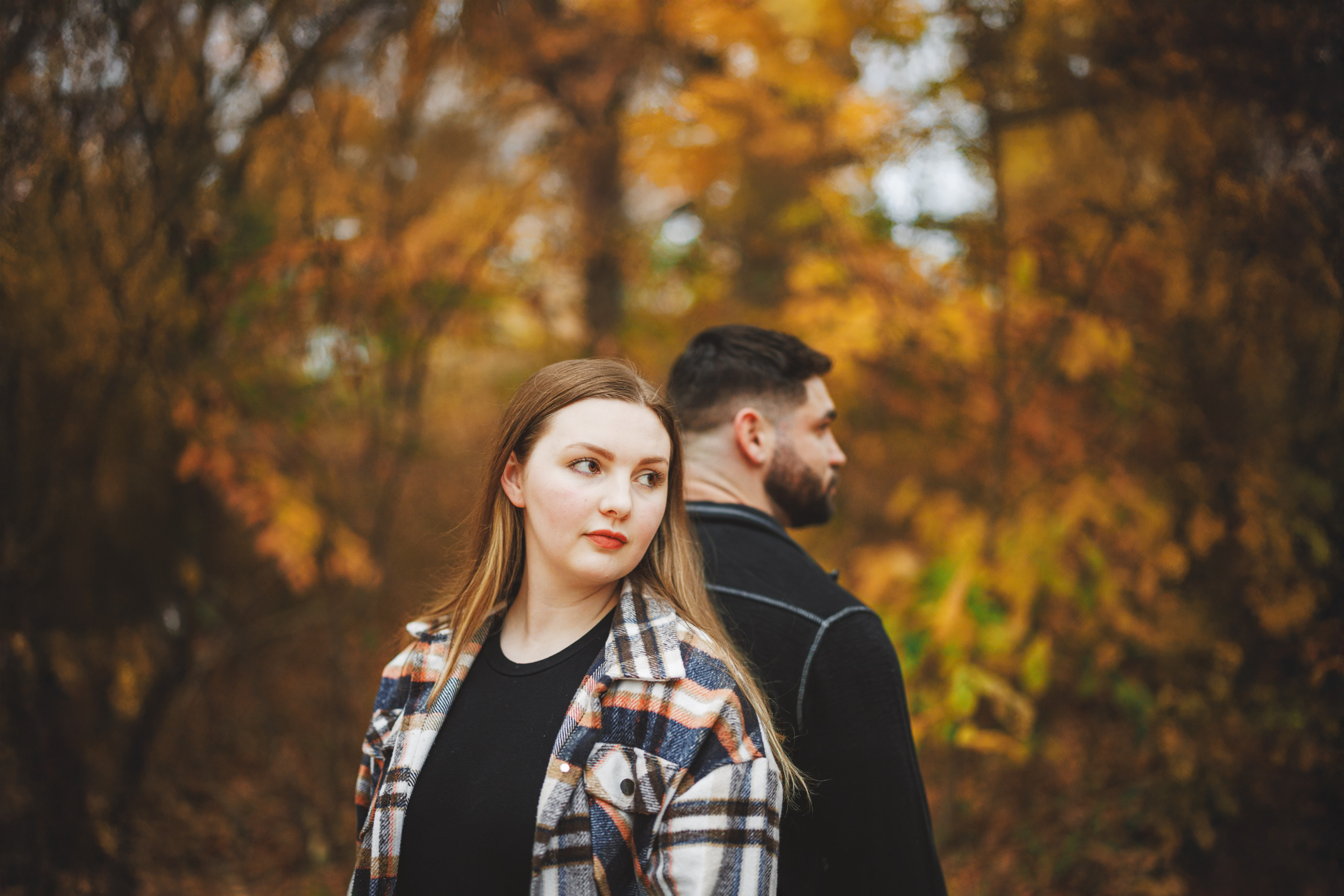 A woman and a man stand back to back in an autumn forest, both looking to the side. The background is filled with trees and fall foliage.