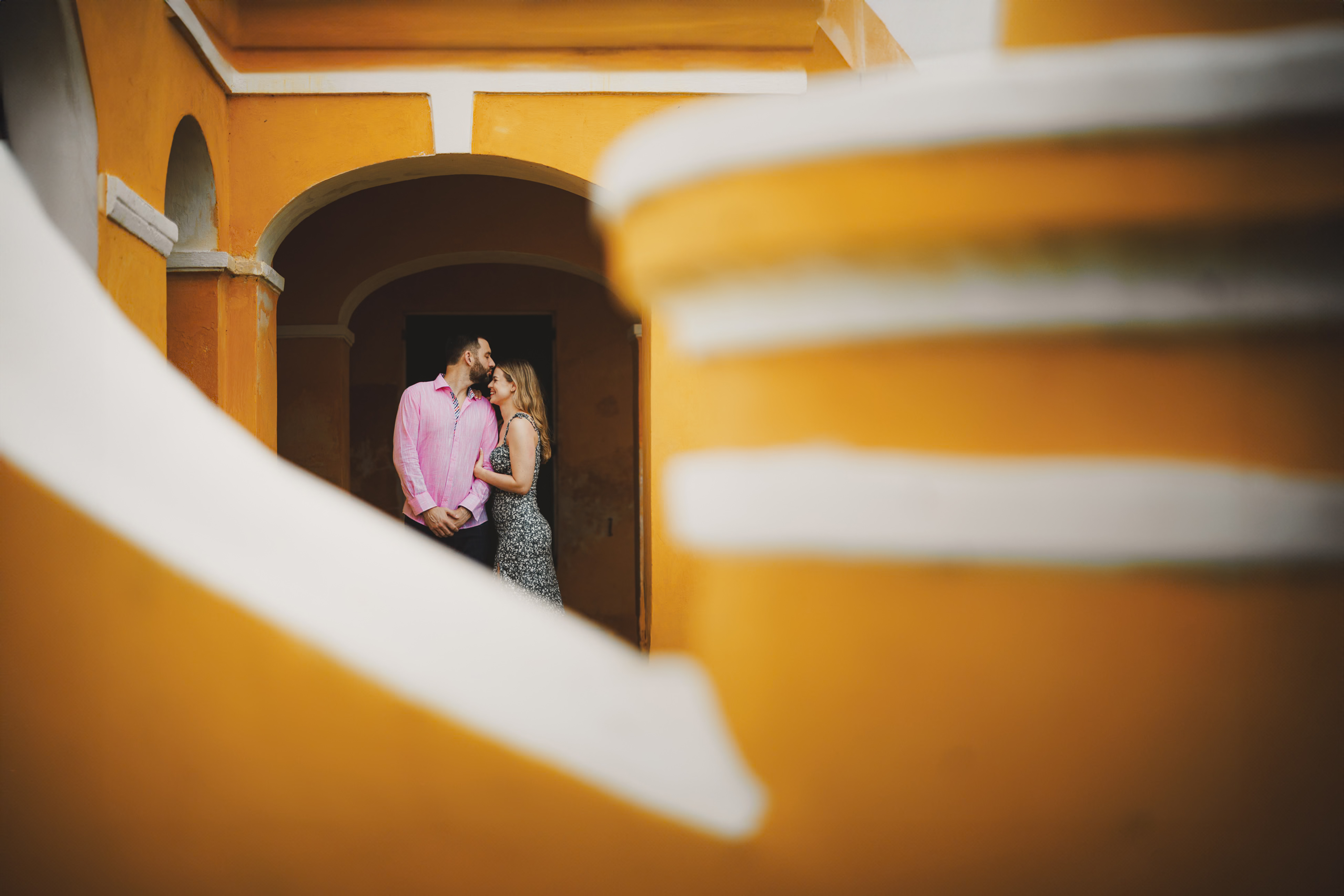Couple standing in front of a bright orange and white architectural background, facing each other affectionately.