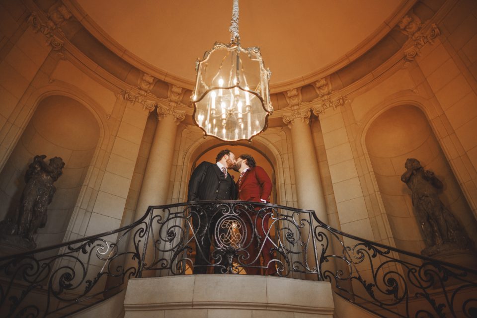 A couple stands on an ornate balcony beneath a grand chandelier in an elegant, circular room with two statues.