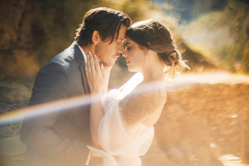 A couple shares an intimate moment outdoors, with soft sunlight and lens flare around them.