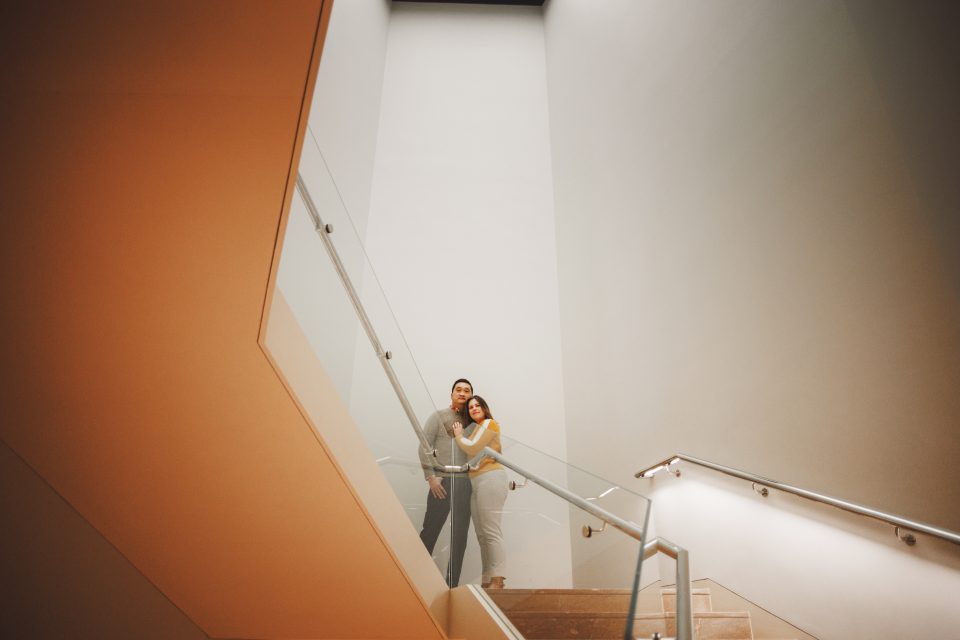 A couple stands on a staircase landing with a minimalist design, featuring glass railings and soft lighting.