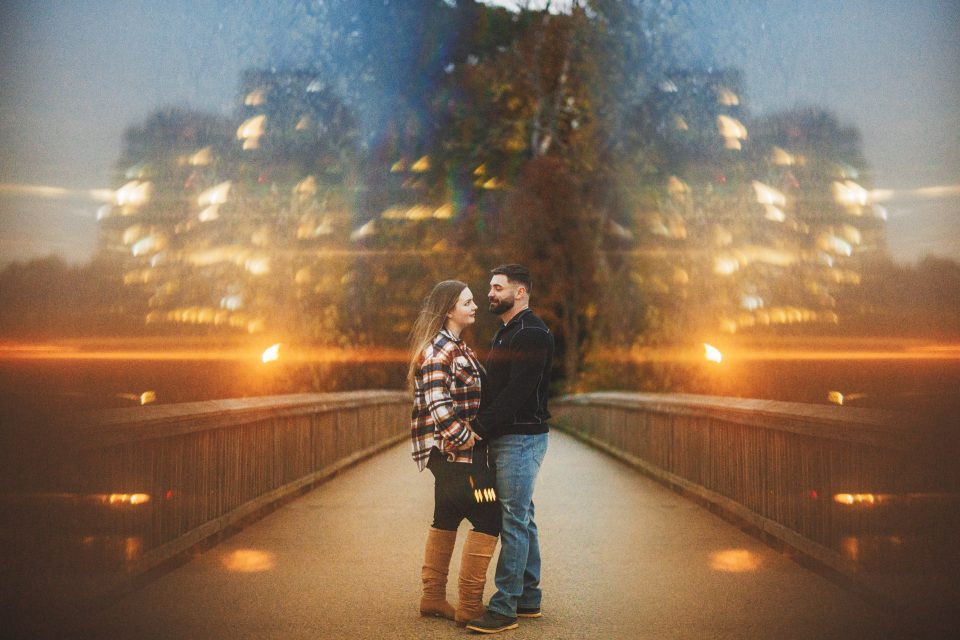 A couple stands on a bridge at dusk, facing each other, with blurred lights and trees in the background, creating a symmetrical reflection effect.