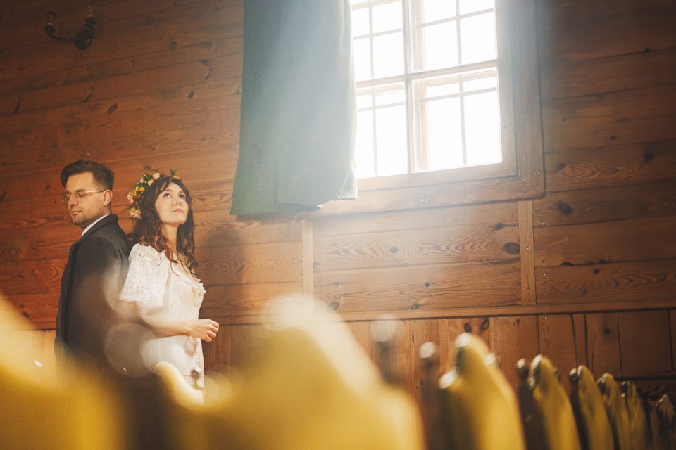 A man and woman wearing a floral crown stand inside a wooden building, near a window with sunlight streaming in.