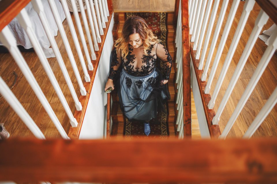 A woman in a black and gray dress descends a wooden staircase, holding the railing.