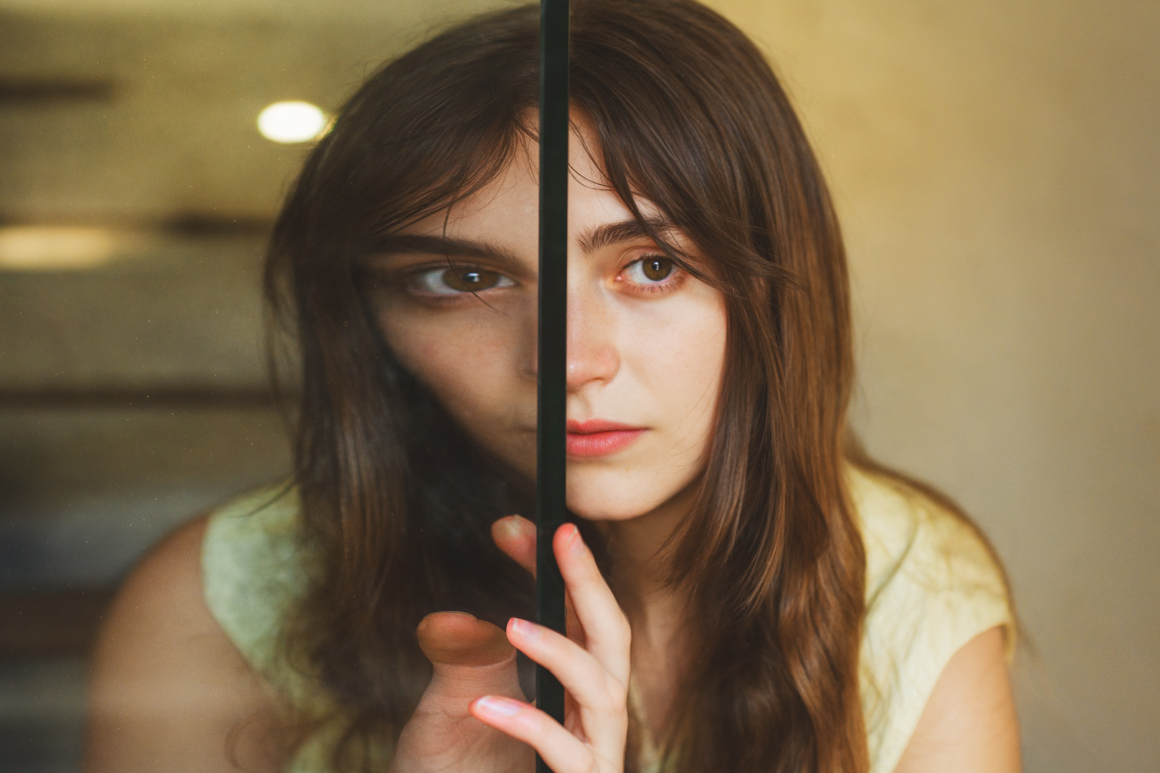 A woman with long brown hair stands behind a glass panel, which distorts part of her face. She gazes forward with a neutral expression.