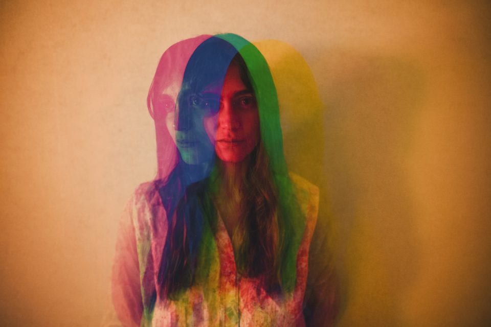 A person stands against a textured wall with a rainbow-colored, triple-exposure effect, creating overlapping silhouettes in red, green, and blue hues.