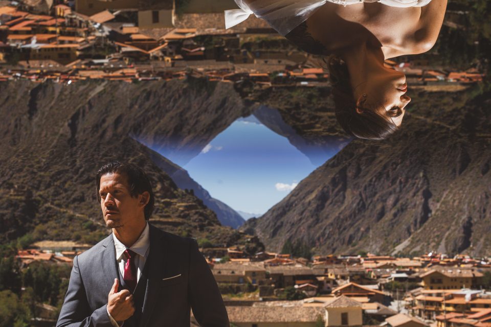 A man in a suit stands in front of a mountainous landscape, with an upside-down image of a woman above him.