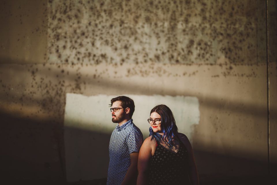 A man and woman standing in front of a textured, sunlit wall. Both wear glasses; the woman has blue-tinted hair.