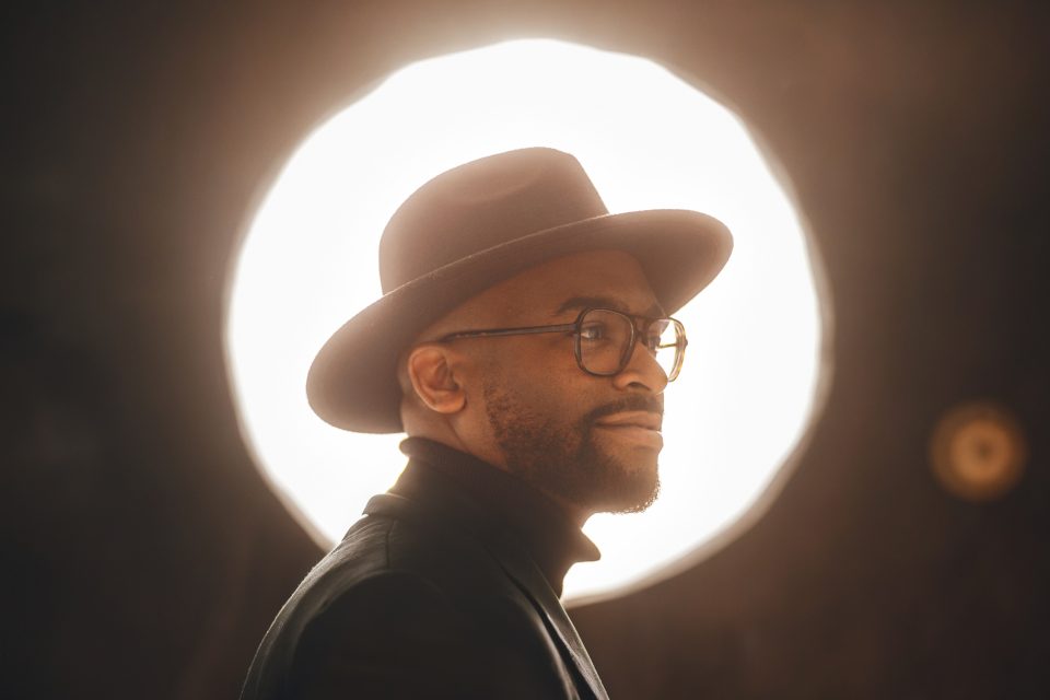 Man in a wide-brimmed hat and glasses looks to the side, standing against a bright circular light in the background.
