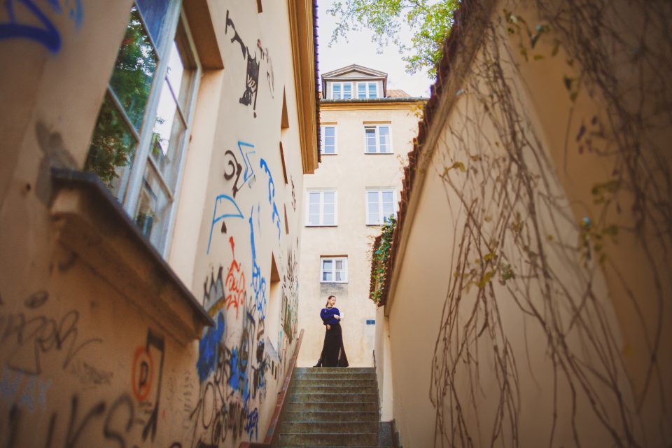 A person stands at the top of a graffiti-lined stairway between two narrow buildings, with vines on the right wall and a multi-story building in view.