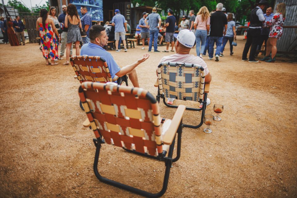 Two people sit on woven lawn chairs facing a group of people gathered in an outdoor area. One person gestures while talking. Drinks are on the ground beside them.