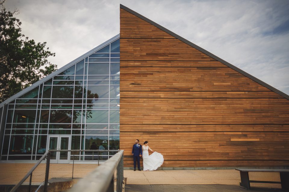 A couple stands in front of a modern building with glass and wooden elements. The bride is in a white dress and the groom in a blue suit.