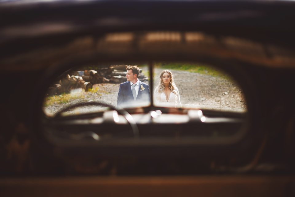 Bride and groom walking outside, viewed through the rear window of a vintage car.
