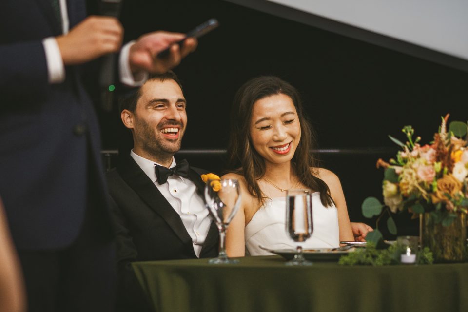A couple in formal attire sits at a table, smiling. A person stands nearby, partially seen, holding a microphone. A floral centerpiece and glassware are on the table.