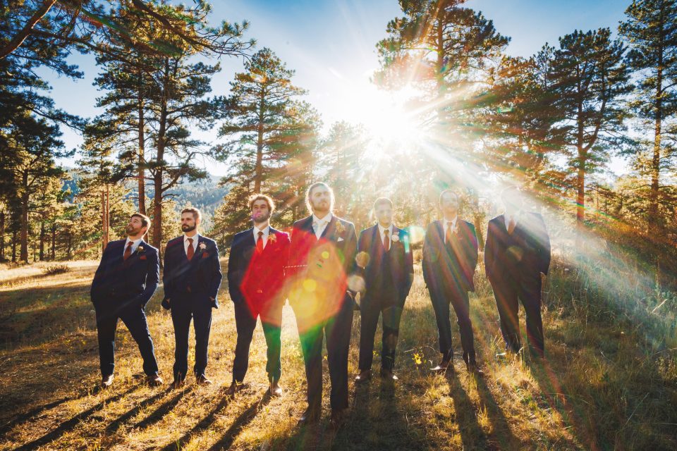 Seven men in suits stand in sunlight in a forested area. Sun rays create a lens flare effect, casting long shadows on the grass.