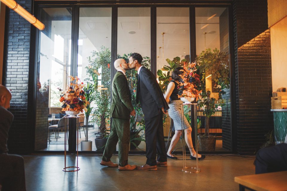 Two men in formal attire share a kiss in front of a glass wall, with plants and flower arrangements nearby.