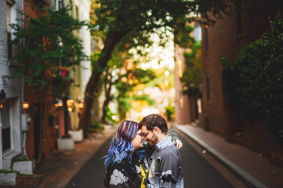 A couple embraces and leans in together on a quiet, tree-lined street. The woman has blue hair and a patterned sweater, and the man has glasses and a patterned shirt.